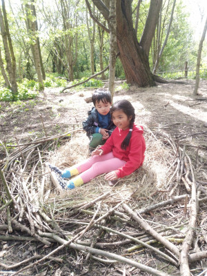 two children with dark hair and skin sat in the woods in a giant pretend nest made from straw material