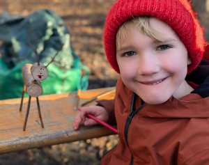 a young child with blonde fringe wearing a coat and a red knitted hat smiling sat next to a reindeer craft made with twigs, the boy is holding a felt tip pen and the reindeer has a hand drawn smiley face on it
