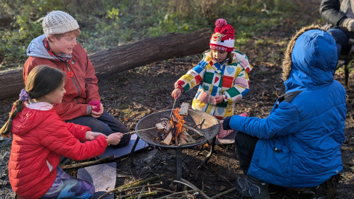 four children wearing coats sat on the floor in a woodland around a firepit toasting marshmallows