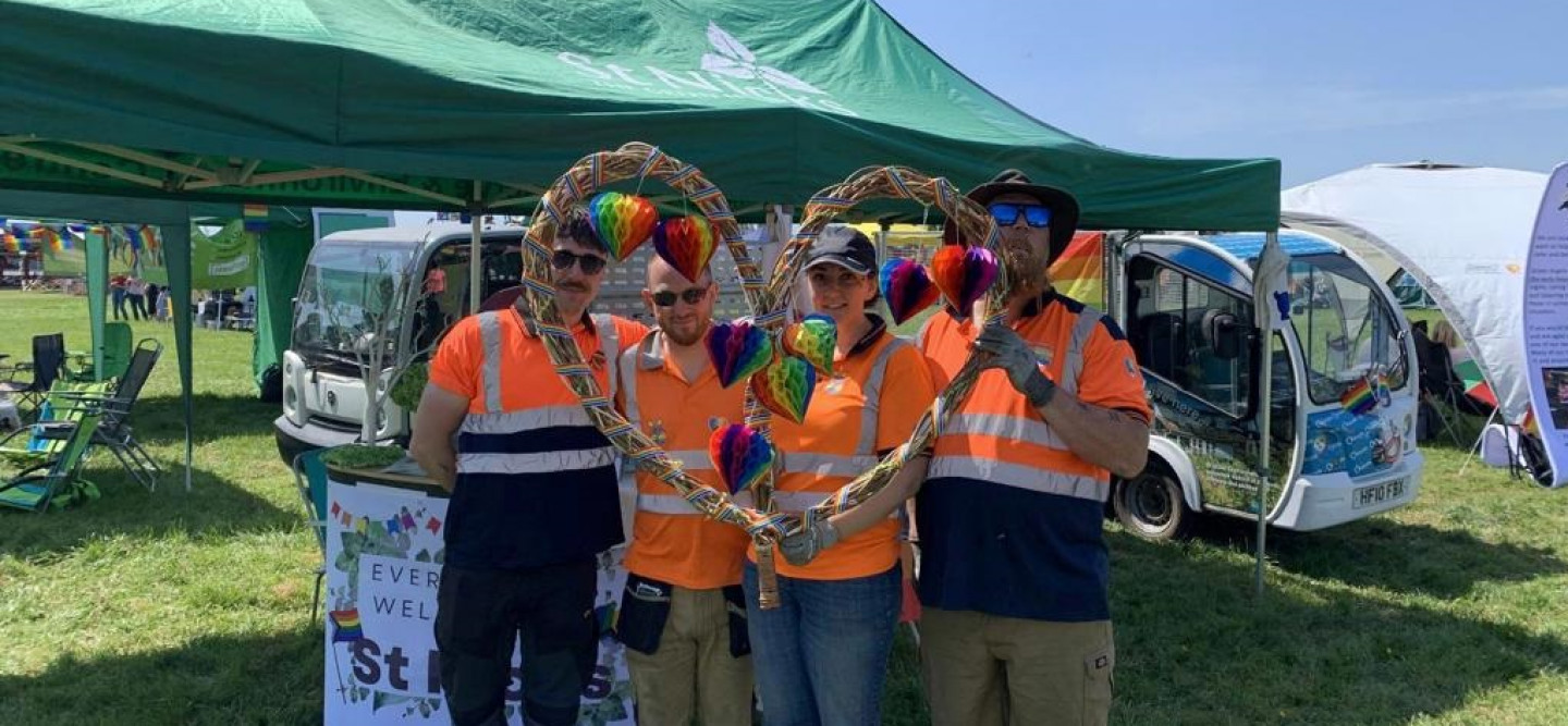 Four smiling people look into the camera three are men and one is a woman. They are holding a woven heart covered in rainbows they are stood in front of a gazebo in a park behind them is a notice that reads everyone is welcome at St Nicks