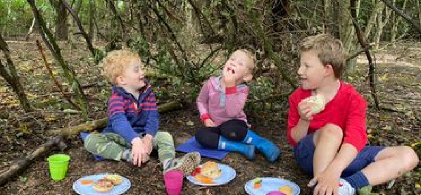 Three children sit on a forest floor eating from plastic plates they are smiling and looking at each other