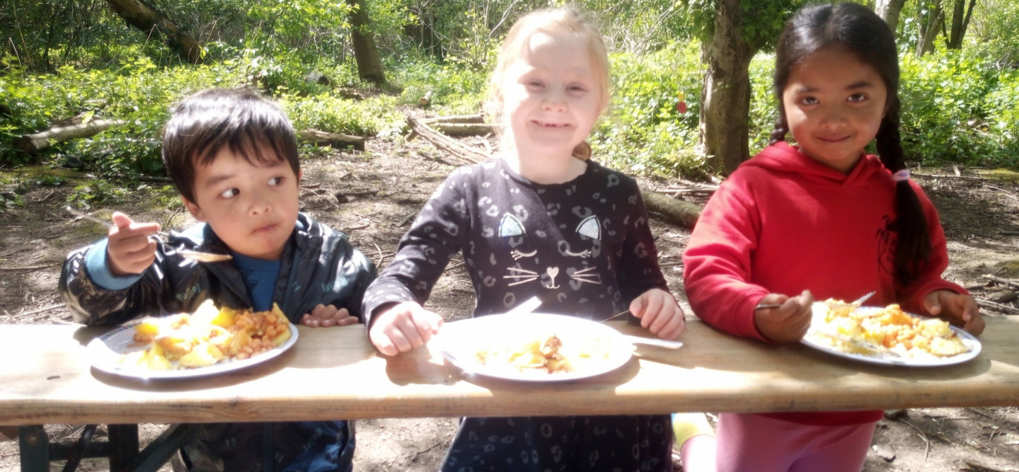 three children, two with dark hair and dark skin and a third with blonde hair and pale skin sit on the floor using a bench as a table. They are eating jacket potatoes with beans. The two girls (one blonde and one dark haired) are smiling at the camera. th