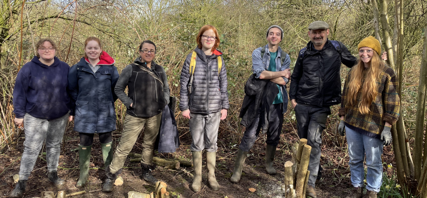 Seven people lined up and smiling for the photo. They are in a woodland and have coppiced stumps of wood at their feet.