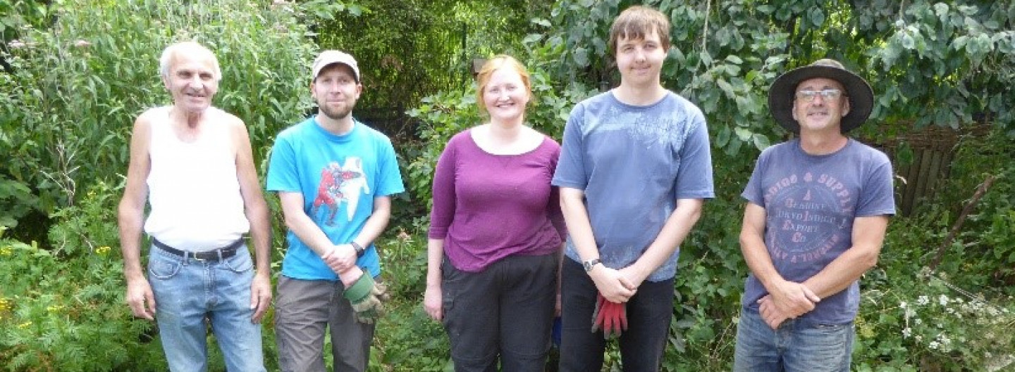 Harriet is a young lady with red hair, she is wearing a bright purple top and black trousers. She is stood with four males of various ages most in t shirts, one is in a white vest. They are all stood in a row behind a bench they have just made. Harriet is