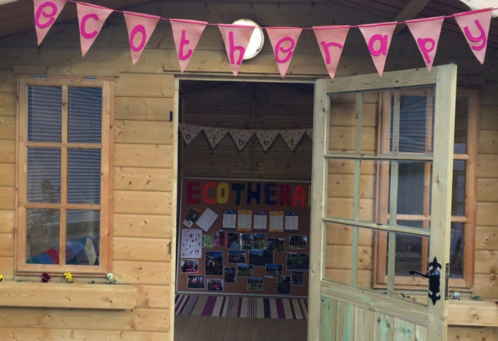 A small wooden shed with open door. There is bunting hanging from an overhand sheltering the door. The bunting is pink and spells out the word ecotherapy.