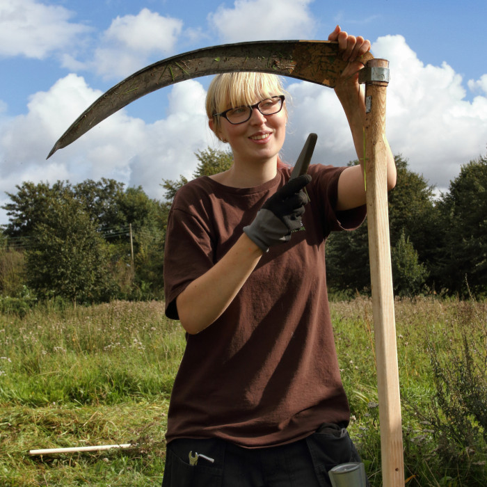 Eithne, a young lady with a long blond fringe touching the frame of her black glasses stands next to a scythe, she is facing the camera and the image shows her from mid thigh her left hand is supporting the top of the scythe and in her gloved right hand s