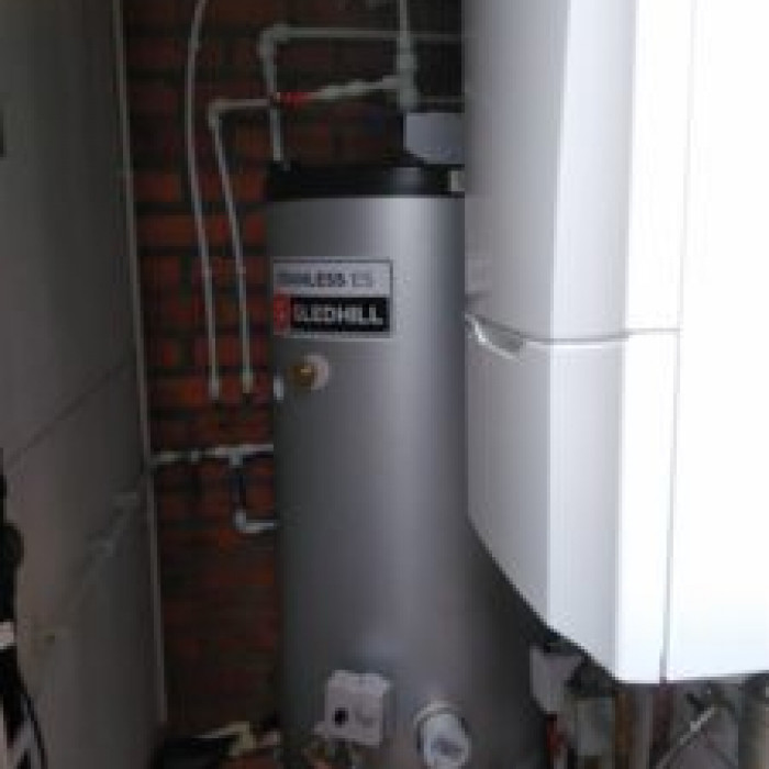 A renovated brick shed containing a water heater and boiler. the boiler is a typical white rectangle. the water heater is a silver tube with lots of pipes mounted on the brickwork above it and coming from the front to the floor.