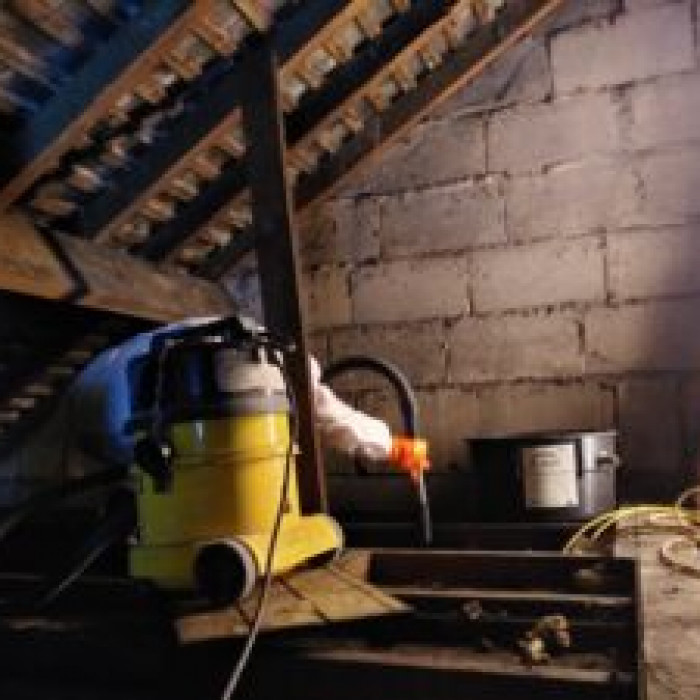 A person in a white safety suit and orange gloves is in a bare brick loft the roof slates and beams are visible the person is using a vacuum between floor joists