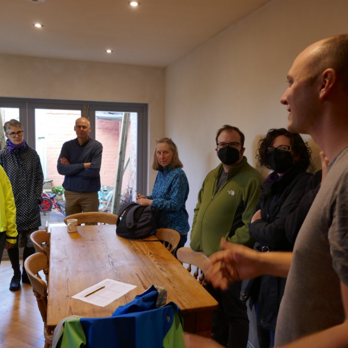 a mixed group of men and women listen to a man talk about his home. They are stood in a semi circle around a dining table with 6 chairs