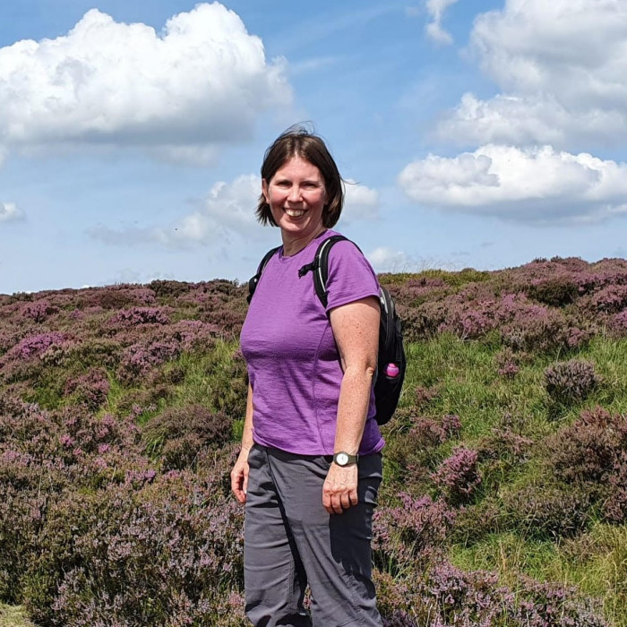 A middle aged white woman with a dark brown bob smiles at the camera. She is wearing hiking gear including a rucksack and is on a moor land surrounded by purple heather