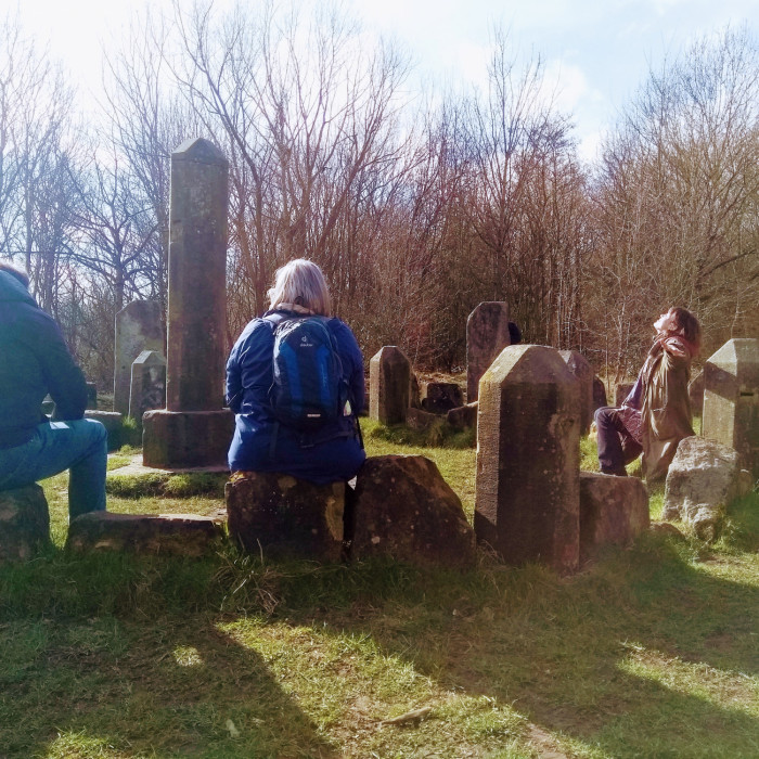 Creative writing participants sit in the sunshine around the St Nicks dragon stones. The participants have their back to the camera. The tutor has arms outstretched and face up to the sun in a joyful pose.