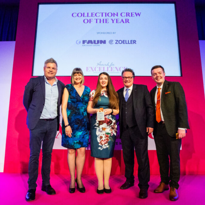 a row of smiling people look down into the camera. There are three men and two women. The men are in suits the women blue and green dresses One of the women is holding an award. The board behind them says 