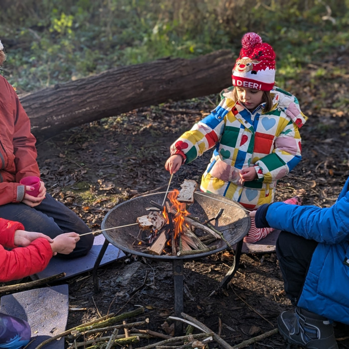 four children wearing coats sat on the floor in a woodland around a firepit toasting marshmallows