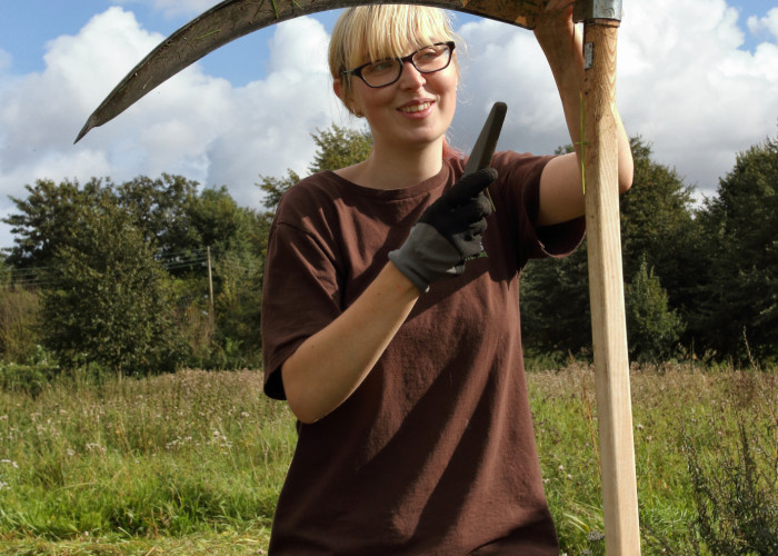 Eithne, a young lady with a long blond fringe touching the frame of her black glasses stands next to a scythe, she is facing the camera and the image shows her from mid thigh her left hand is supporting the top of the scythe and in her gloved right hand s