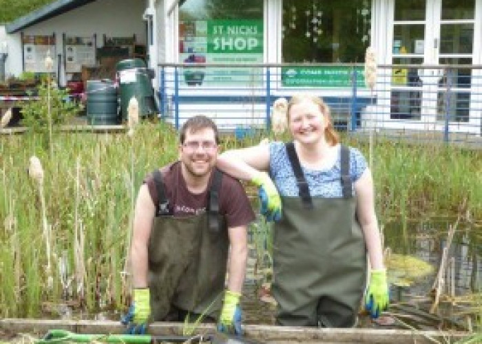 Park Rangers Nick and Harriet both wearing waders and gloves stand in the pond and smiling. Nick is on the left and leaning forwards holding the edge of our pond dipping platform, he is a young, white male with stubble and glasses. Harriet has her elbow o