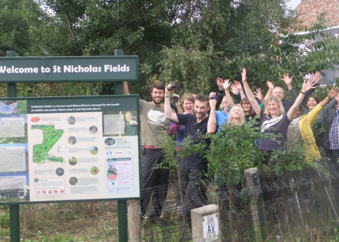 A large group of smiling volunteers cheering with their arms up stand behind a green sign with the banner saying Welcome to St Nicholas Fields