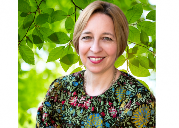 A middle aged white woman with pinky red lipstick and a short soft blonde bob smiles at the camera. She is wearing a thick gold chain necklace and a very colourful floral patterned top in front of a green leafy background