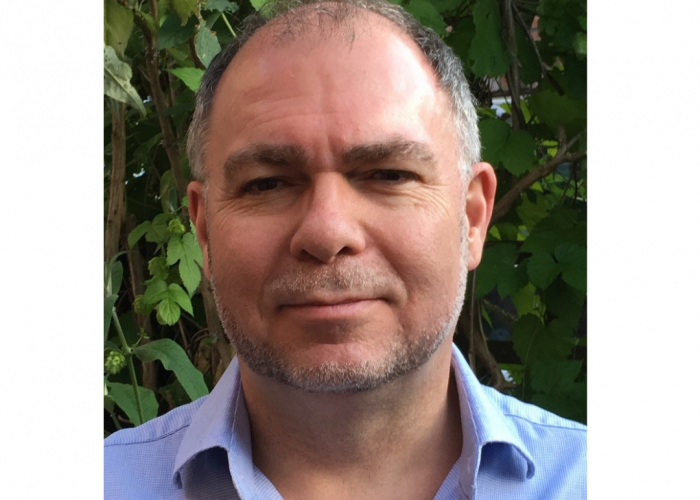 Jamie is a white male with grey stubble, balding on top with areas of dark hair and white hair. He is slightly smiling at the camera wearing a blue shirt. He is stood in front of greenery.