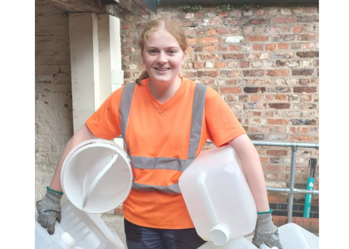 Martha is a young lady with her light brown hair tied back. She is wearing an orange hi vis t shirt and gloves and carries lots of very large catering plastic bottles under her arms