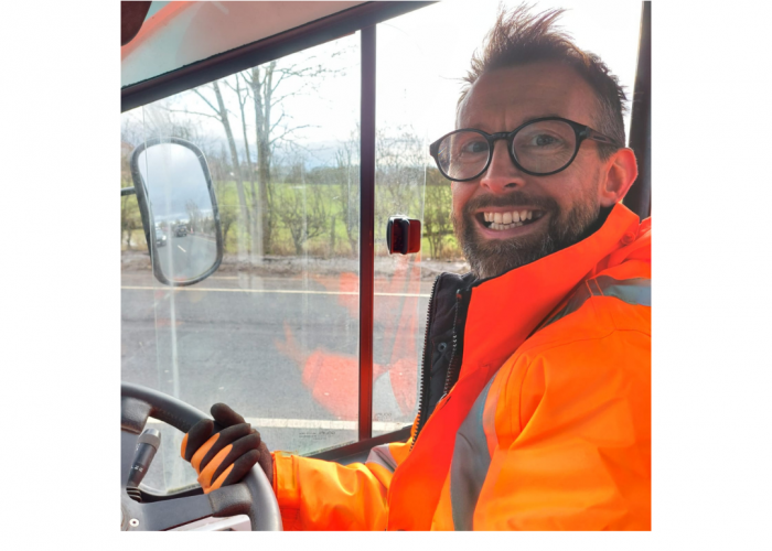 A grinning male with spiky dark hair, large round dark glasses and a bear looks straight into the camera. He is holding a steering wheel and there is an open vehicle window behind him