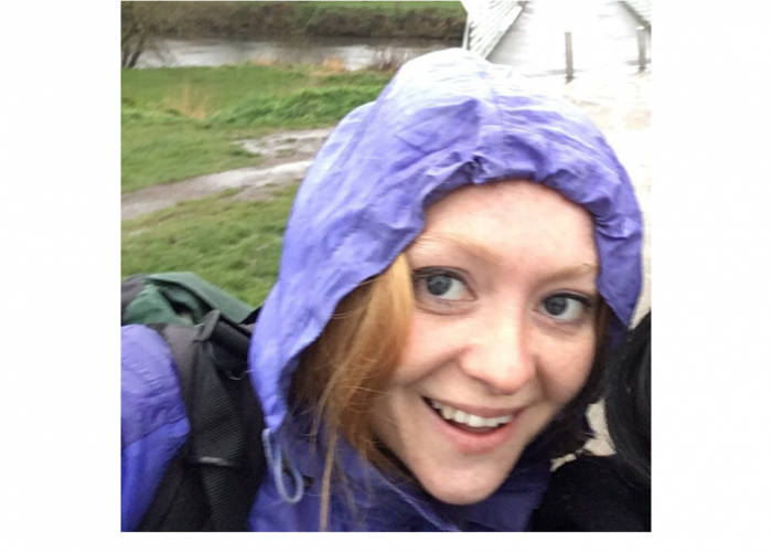 Maria Gill is a young red haired freckled woman. Her hair is incredibly long though it is tucked up into a rain hood. there is a very wet grass floor behind her, a bridge and a river.