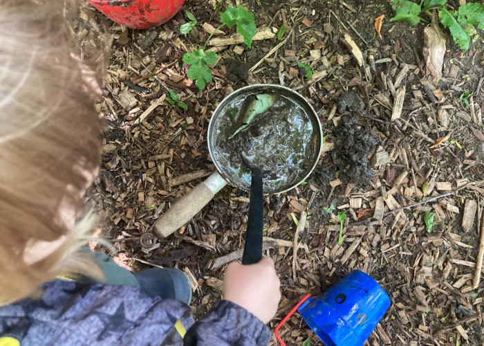 a view from above a child who is stirring a metal pot with a plastic ladle. The pot is filled with water and mud