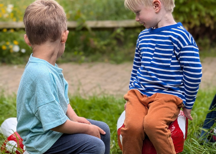 Two small boys sit looking behind them (away from the camera) into the grass. they are sat on bright red stools shaped like mushrooms carved from tree stumps.