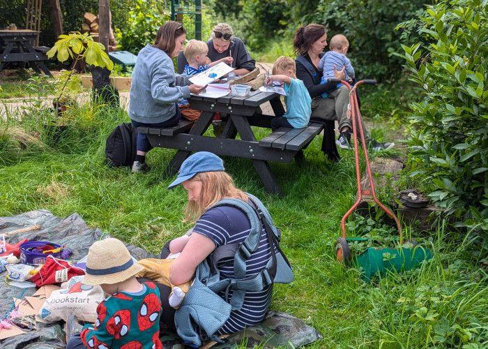 A group of women and young children, mainly toddlers sat in groups, some are on a picnic bench painting, others are sat on the floor using scissors and fabric. they are all outside on grass in a garden.