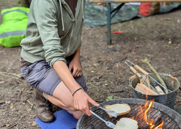 a young curly haired woman in shorts kneels next to a fire pit using tongs to turn pieces of bread