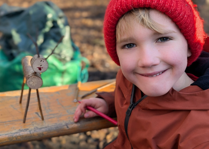 a young child with blonde fringe wearing a coat and a red knitted hat smiling sat next to a reindeer craft made with twigs, the boy is holding a felt tip pen and the reindeer has a hand drawn smiley face on it