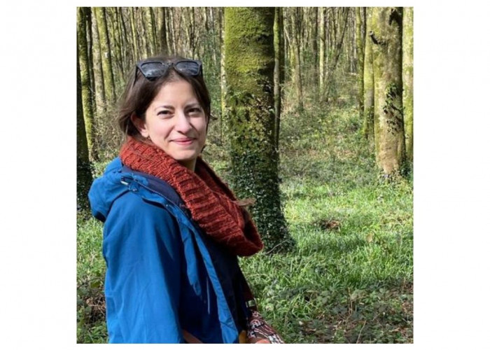 Marina in a woodland wearing a blue jacket and a red scarf, she has sunglasses on her head and is smiling at the camera