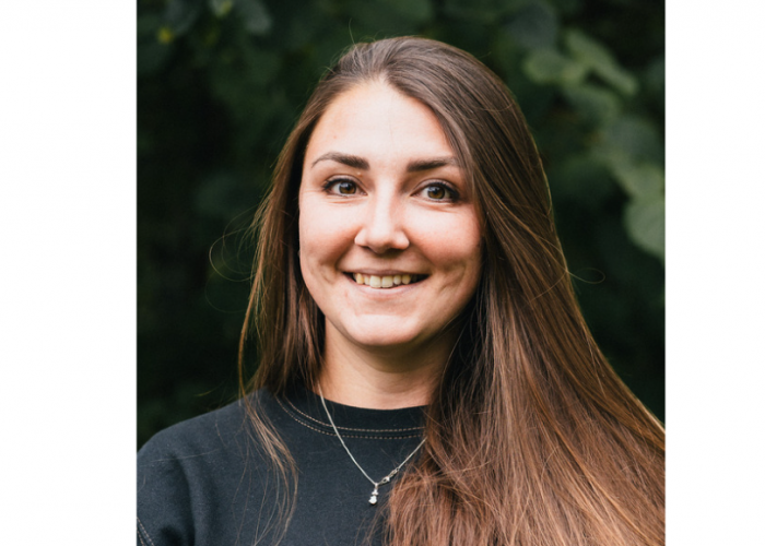 Sam is a young woman with very long, straight brown hair. She is wearing eyeliner and mascara and a necklace over the top of her jumper. She is smiling at the camera