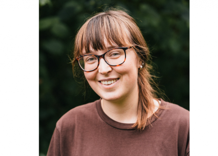 Eithne, a young woman with brown hair tied back from her face, she has a fringe, two ear piercings visible and toroishell glasses. She is smiling widely at the camer