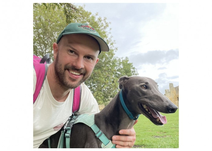 A man in a green baseball cap and a red backpack is smiling at the camera with his arm around a blue greyhound