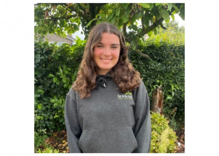 A woman is standing in front of a hedge and flowery plans and smiling at the camera. She has long brown hair and is wearing a grey hoodie with a St Nicks logo on it.