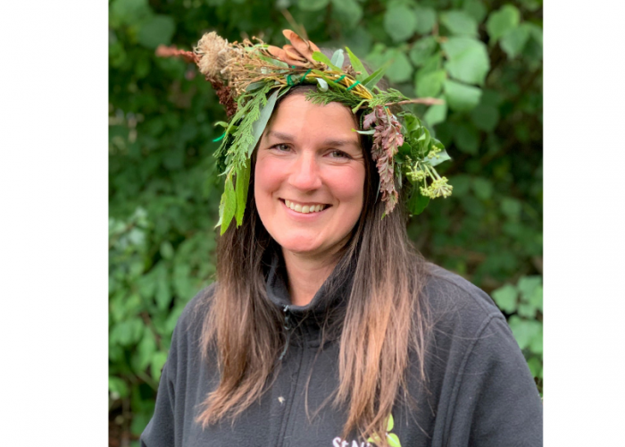 Nicola Ward is a woman with long dark hair, she is smiling at the camera and wears a crown she has made out of green white and brown leaves and flowers on her head