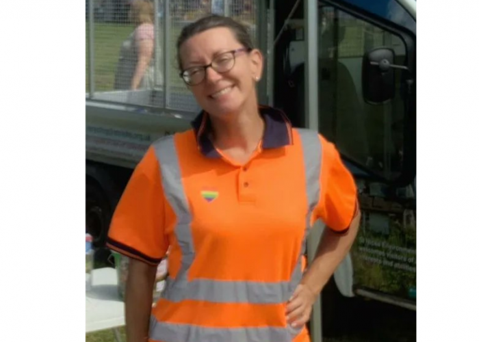 Charlotte Hanson is woman with dark hair tied in a bun at the back of her head she is wearing an orange hi vis top and glasses and is smiling proudly at the camera with her hand on her hip