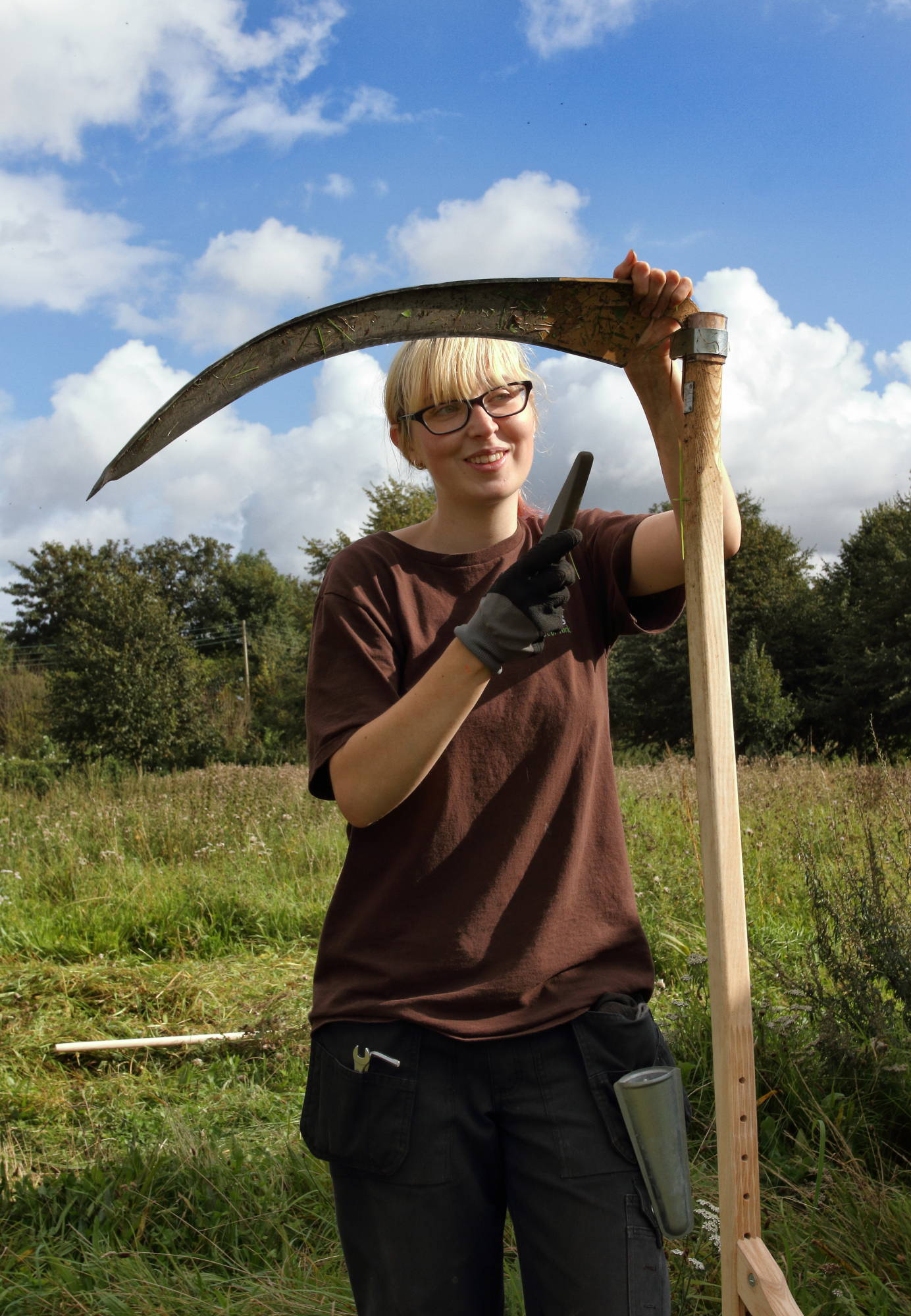 Eithne, a young lady with a long blond fringe touching the frame of her black glasses stands next to a scythe, she is facing the camera and the image shows her from mid thigh her left hand is supporting the top of the scythe and in her gloved right hand s