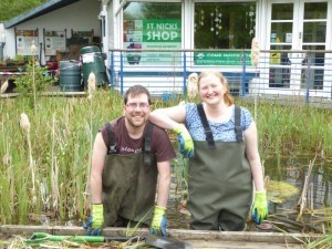 Park Rangers Nick and Harriet both wearing waders and gloves stand in the pond and smiling. Nick is on the left and leaning forwards holding the edge of our pond dipping platform, he is a young, white male with stubble and glasses. Harriet has her elbow o