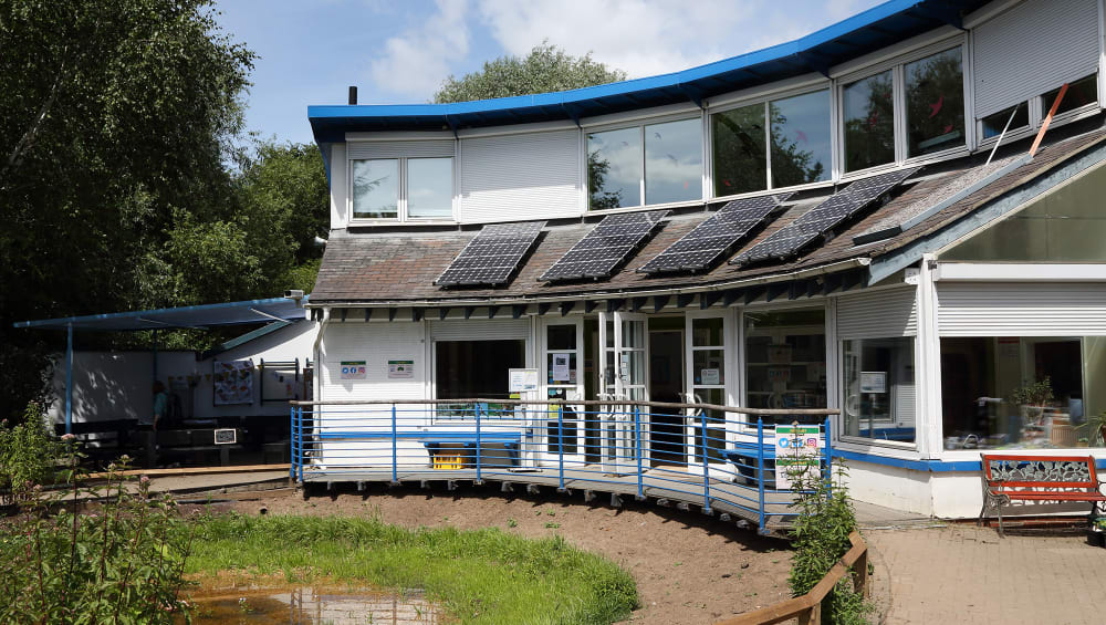 A photo of the St Nicks environment centre, it's a curved white building with blue trimings, solar panels a large pond and benches