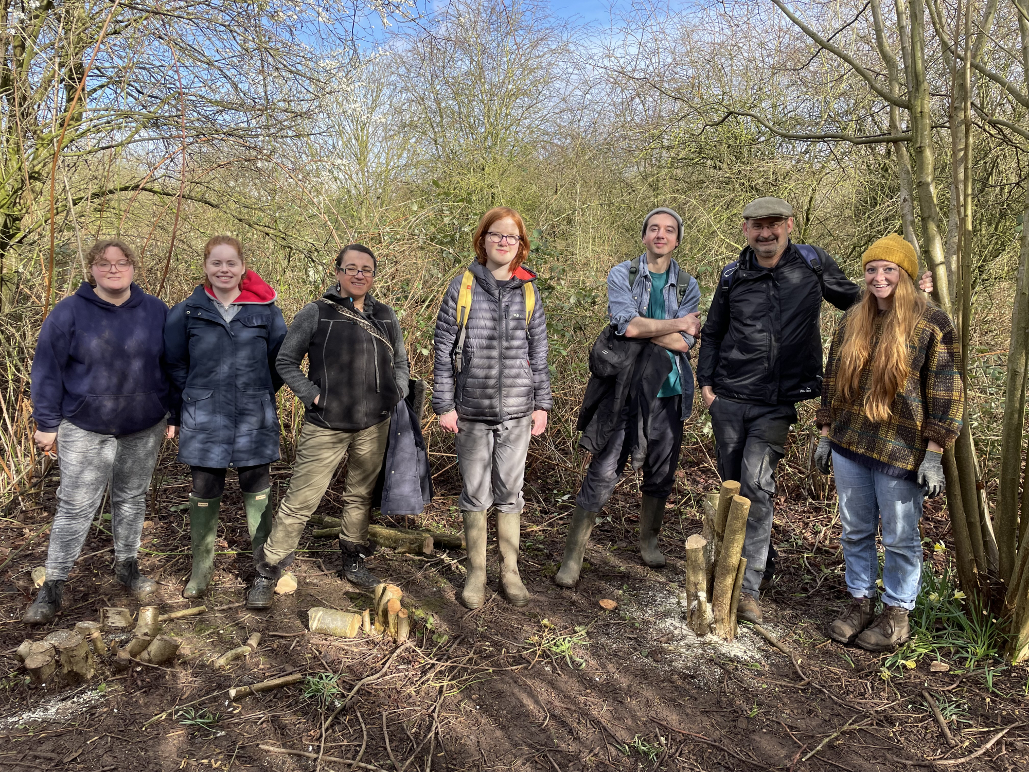 Seven people lined up and smiling for the photo. They are in a woodland and have coppiced stumps of wood at their feet.